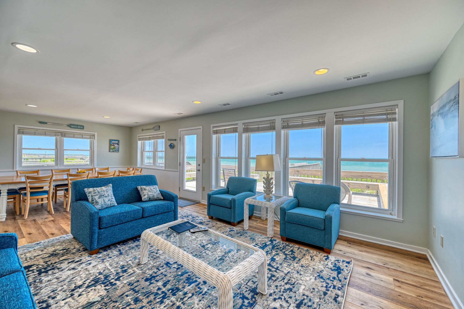 A spacious living room with a view of the beach, featuring blue sofas, a glass coffee table, and large windows.