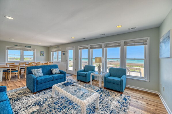 A spacious living room with a view of the beach, featuring blue sofas, a glass coffee table, and large windows.