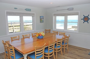Uma sala de jantar com mesa e cadeiras de madeira, grandes janelas com vista para o oceano e decoração náutica.