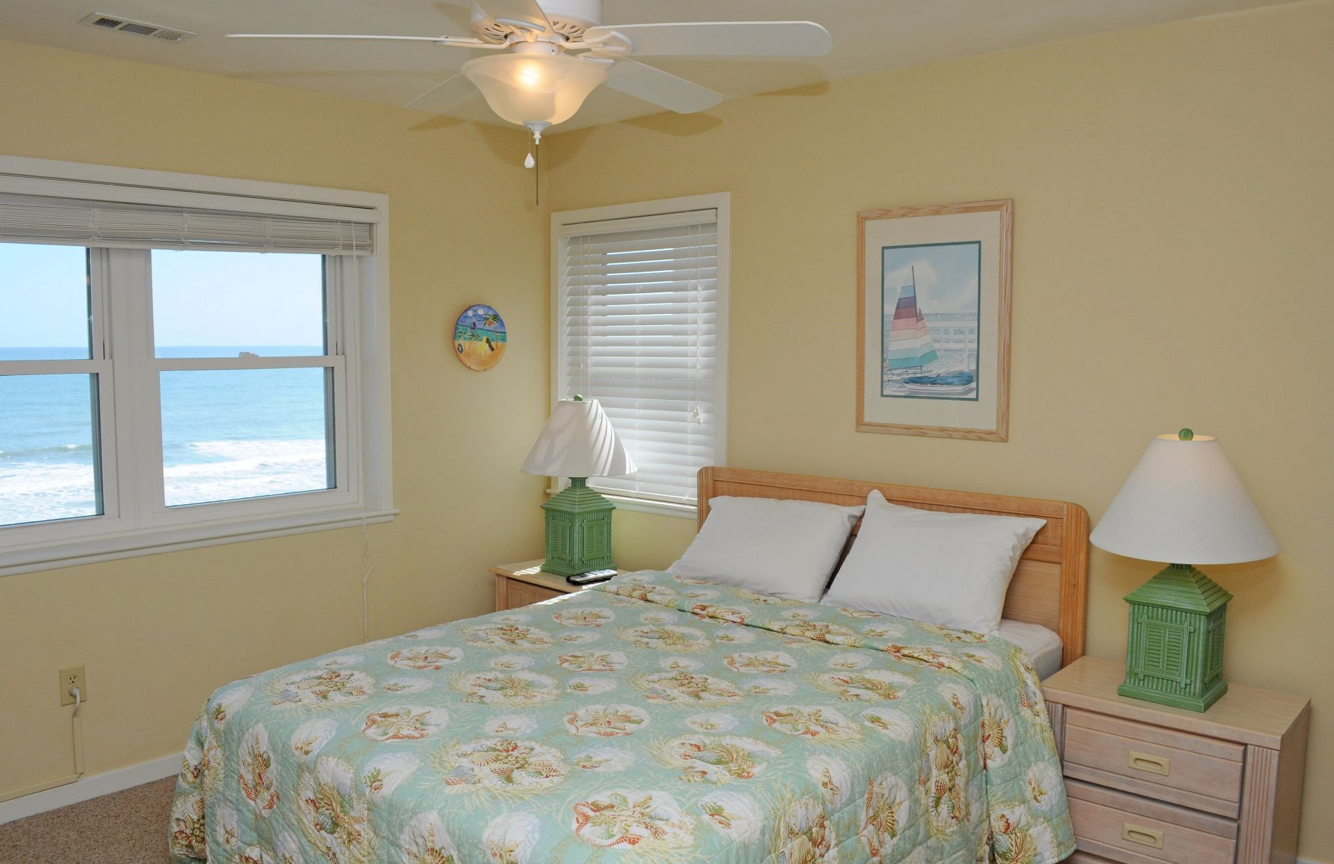 A bedroom with a bed, two nightstands, a ceiling fan, a window with a view of the ocean, and a framed picture on the wall.