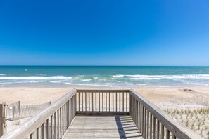 Ein hölzerner Steg führt zu einem Strand mit Wellen, blauem Himmel und sandigem Ufer.
