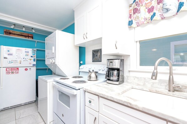A kitchen with white cabinets, a stove, a coffee maker, and a window with a view. - Rhyne-O - Cozy & Comfortable Beach Cottage!!! (Surf City)