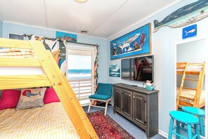 A room with a bunk bed, a desk, a chair, a TV, and a large window with a view of the ocean.