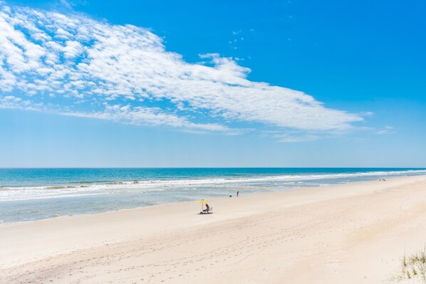A beach with a clear sky and a few clouds, a sandy shore, and a person sitting under an umbrella.