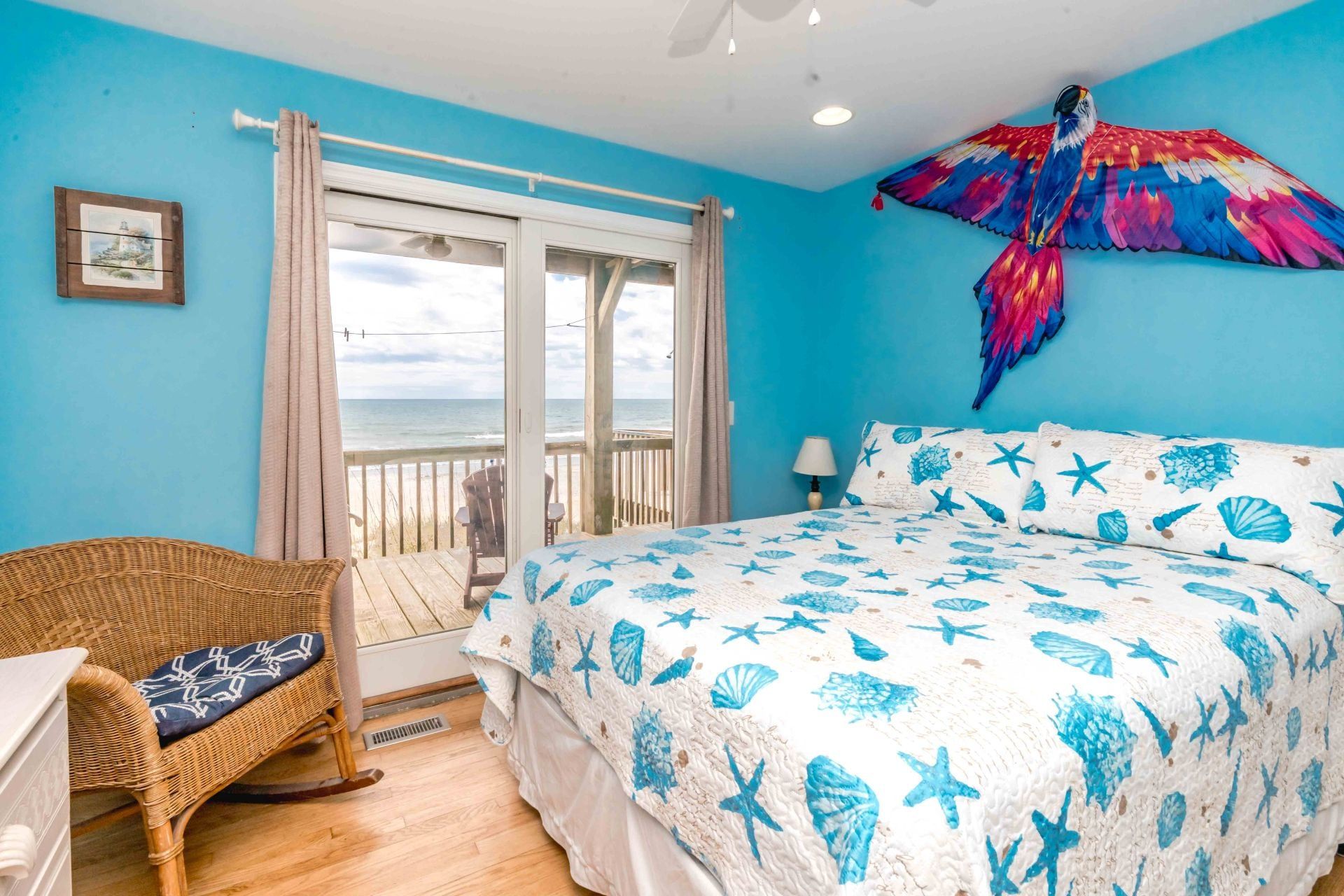 A bedroom with a bed, a wicker chair, a view of the ocean, and a colorful bird decoration.