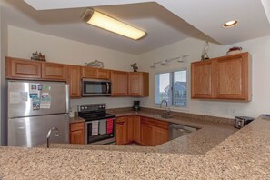 A kitchen with wooden cabinets, a stainless steel refrigerator, a microwave, an oven, granite countertops, and a window with blinds.