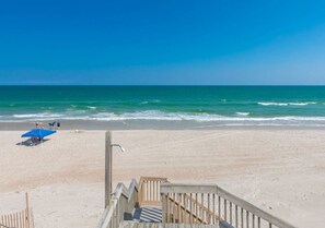 A beach with a wooden boardwalk leading to the water, a blue umbrella, and a few people in the distance.