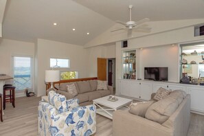 A spacious living room with a high ceiling, a ceiling fan, a television, and a kitchen area with a glass display cabinet.