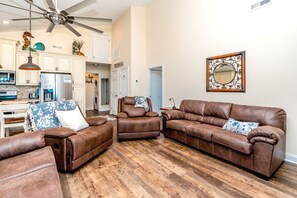 A spacious living room with a brown leather sofa set, a ceiling fan, and a kitchen in the background.