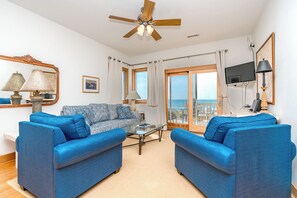 A living room with a blue sofa set, a ceiling fan, a television, and a view of the sea through large windows.