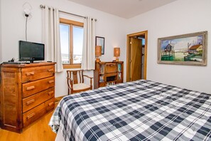 A bedroom with a bed, wooden dresser, chair, desk, lamp, painting, and a window with curtains.