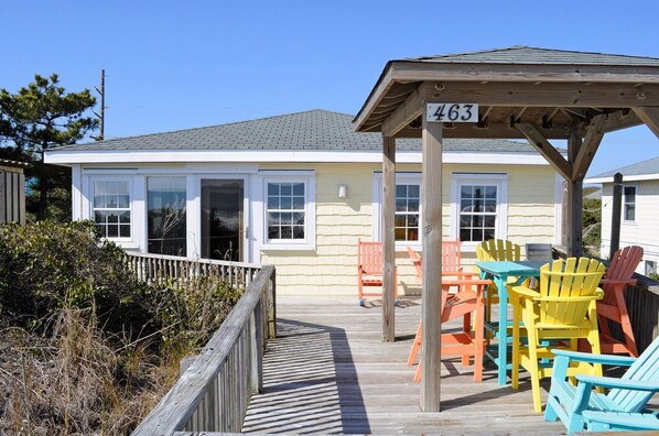 A wooden deck leading to a house with numbered windows, outdoor seating with colorful chairs, and a clear sky. - Tumble Inn - Oceanfront home in Topsail Beach! Gazebo & Dog Friendly! (Topsail Beach)