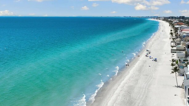 Ein Küstenstrand mit klarem türkisblauem Wasser, einem Sandufer und Gebäuden entlang der Küste.