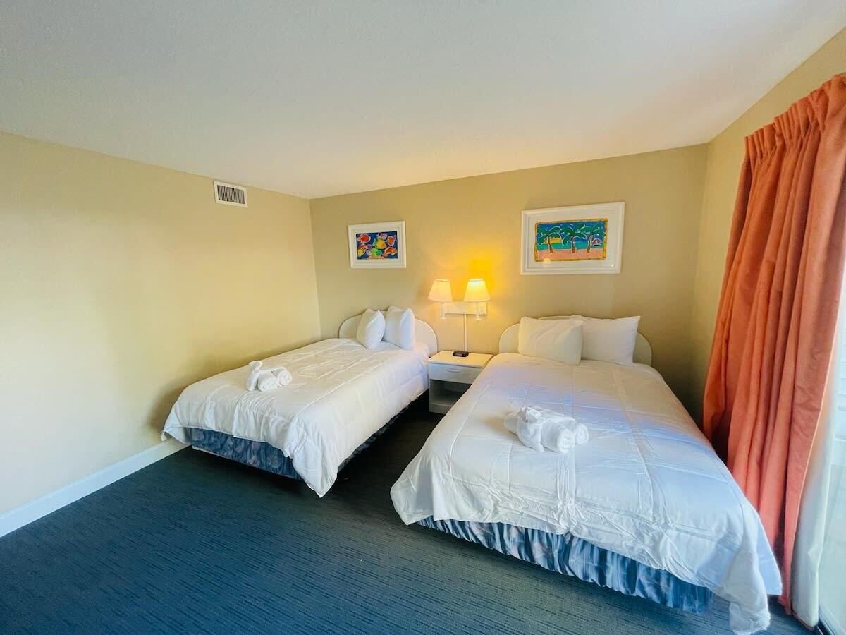 A hotel room with two single beds, a nightstand with a lamp, and two framed pictures on the wall.