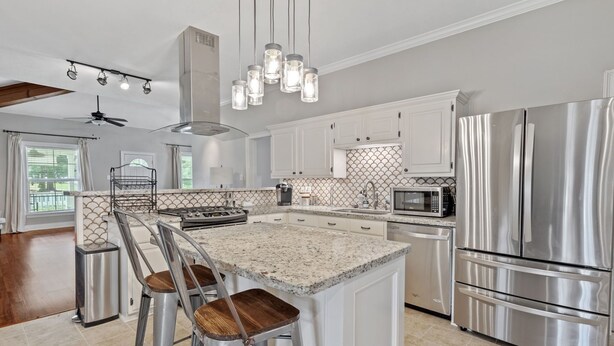 A modern kitchen with white cabinetry, a stainless steel refrigerator, a granite island with bar stools, and pendant lighting.