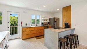A modern kitchen with a central island, wooden cabinets, and a built-in oven. There are bar stools and a dining table with chairs. - Modern Coastal Home, Close to Beach with Pool, Spa (Kitty Hawk)