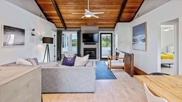 A modern living room with a sofa, a dining table, a ceiling fan, and a fireplace. - Modern Coastal Home, Close to Beach with Pool, Spa (Kitty Hawk)