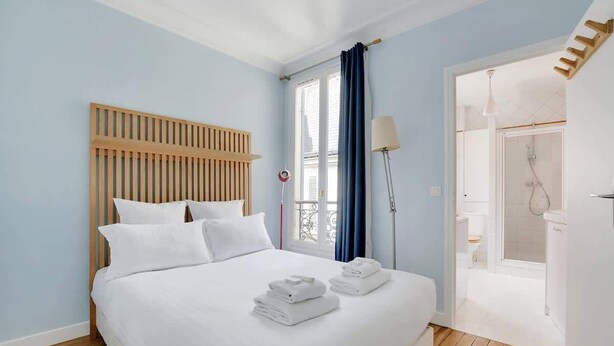 A hotel room with a wooden headboard, a bed with white linens, a bathroom with a shower, and a balcony with a blue curtain. - An Amazing 2-BR/1BA in Ternes - Arc de Triomphe (Paris)