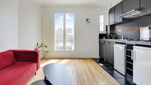 A modern apartment with a red sofa, a dining table, a kitchen with black cabinets, and a large window providing a city view. - An Amazing 2-BR/1BA in Ternes - Arc de Triomphe (Paris)