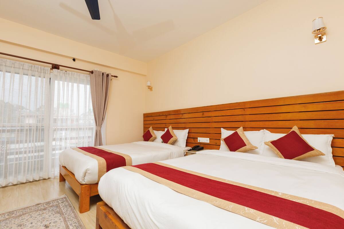A hotel room with two beds, wooden headboard, white bedding, red and beige accents, a wooden floor, a window with sheer curtains, and a wall-mounted light fixture.