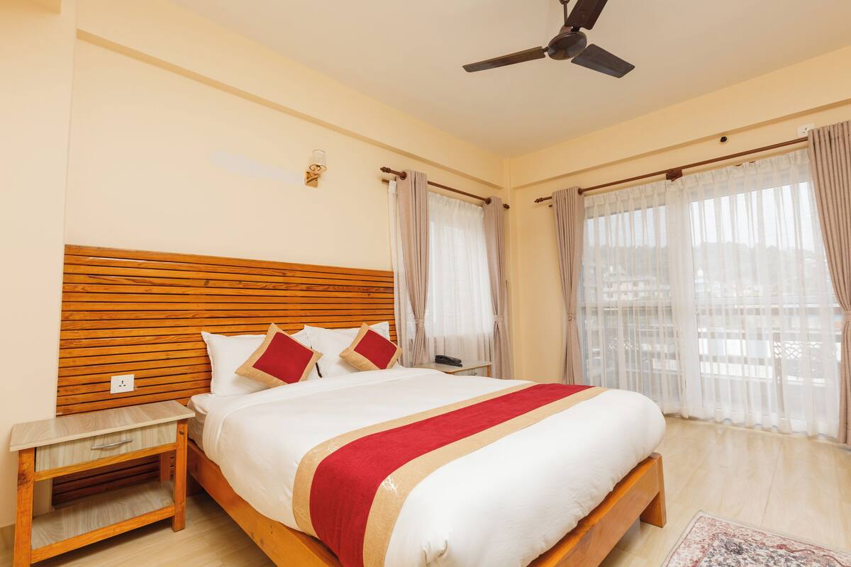 A hotel room with a wooden headboard, a bed with white bedding and a red accent, a nightstand with a drawer, a ceiling fan, and large windows with sheer curtains.
