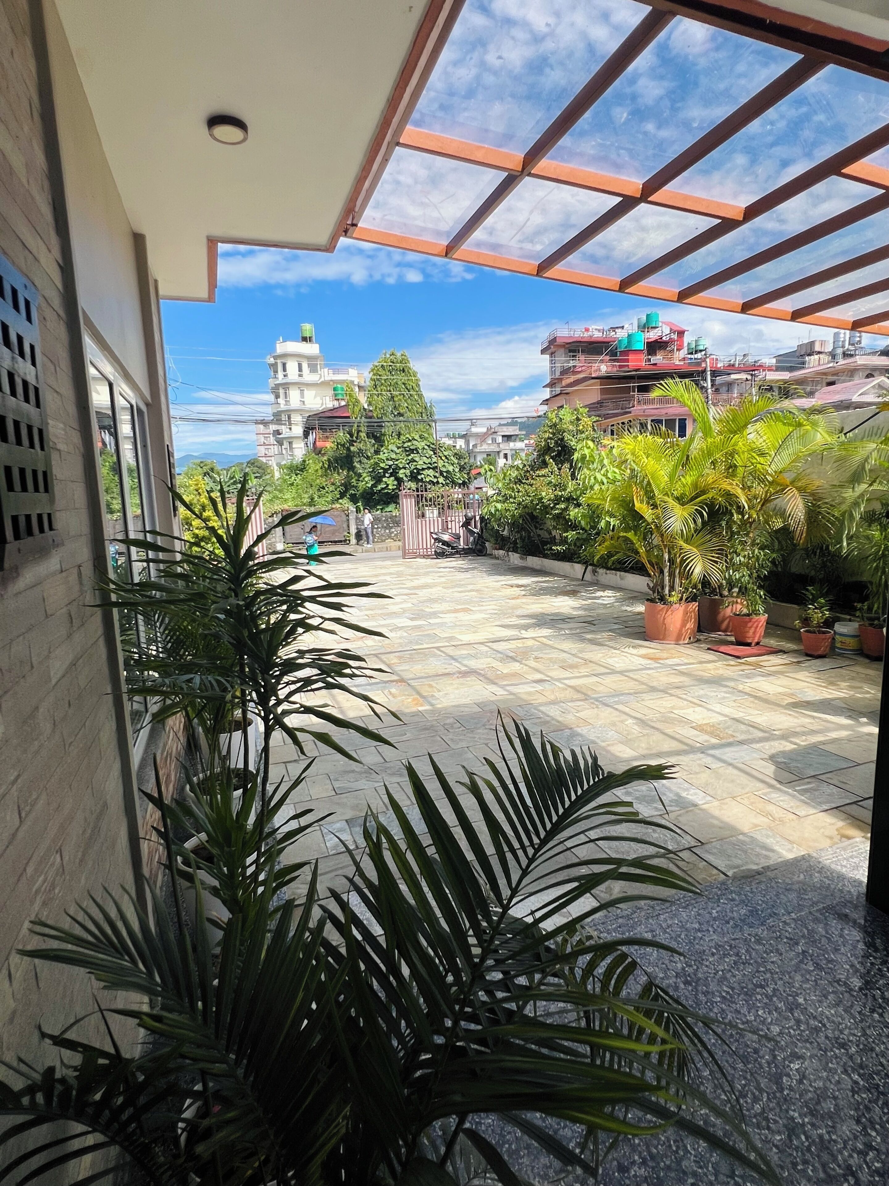 Un espacio al aire libre techado con plantas en macetas, piso de baldosas y vista de edificios circundantes.