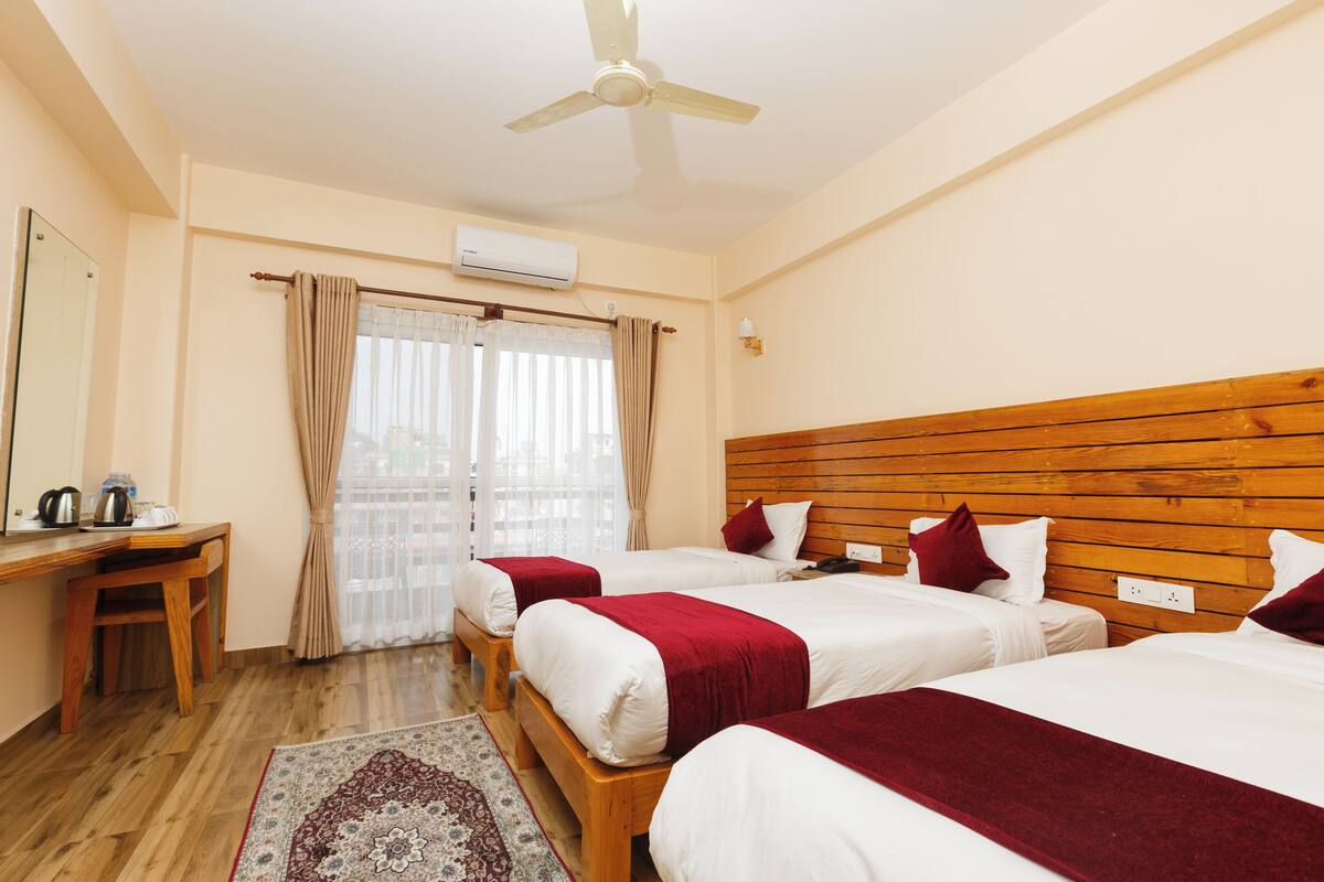 A hotel room with three beds, a wooden desk, a chair, a ceiling fan, and a window with curtains.