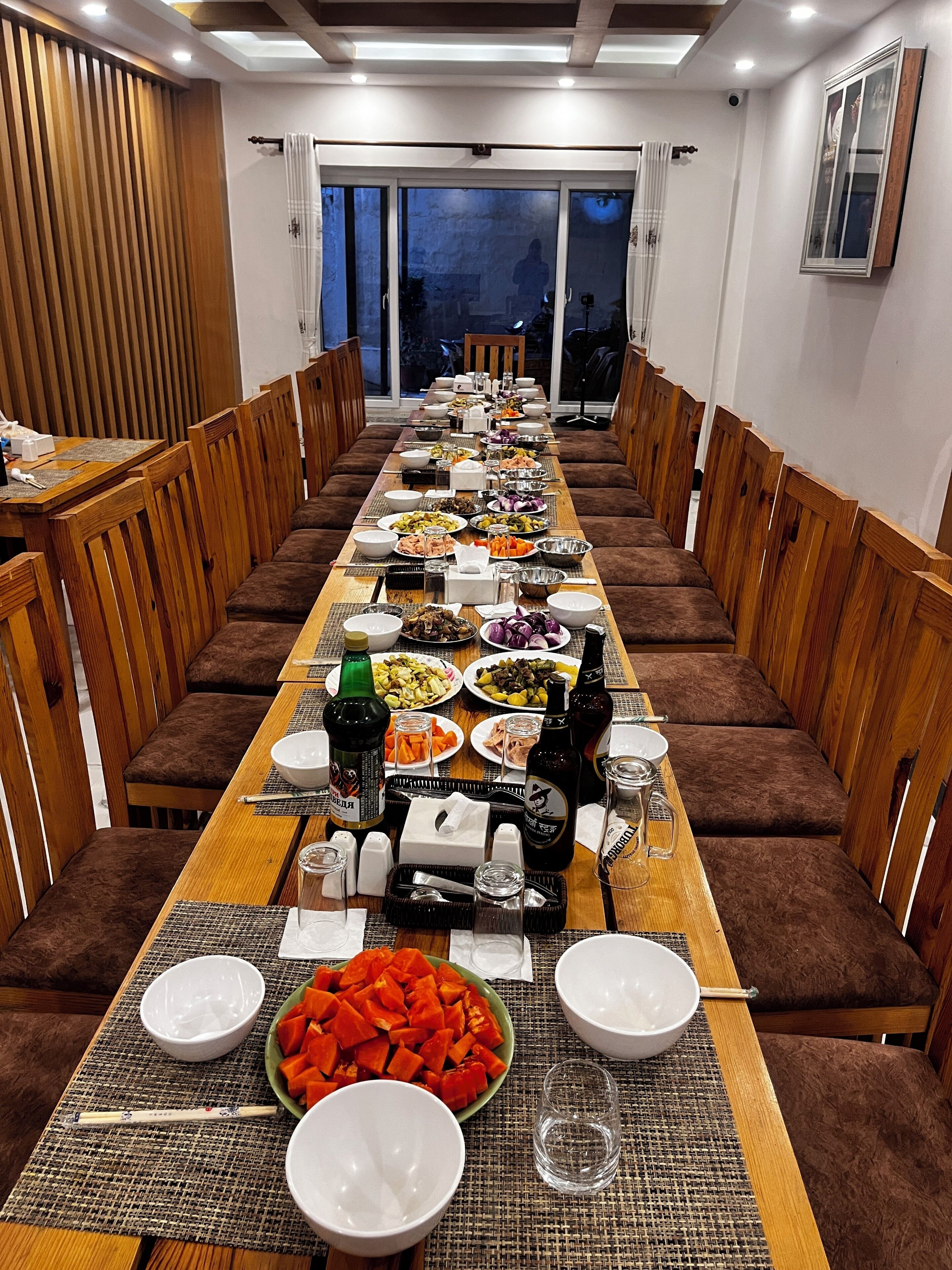 Una larga mesa de comedor dispuesta con diferentes platos, vasos y botellas, rodeada de sillas de madera.