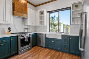 Une cuisine moderne avec des meubles blancs et turquoise, des appareils en acier inoxydable et une grande fenêtre donnant sur des arbres et des immeubles.