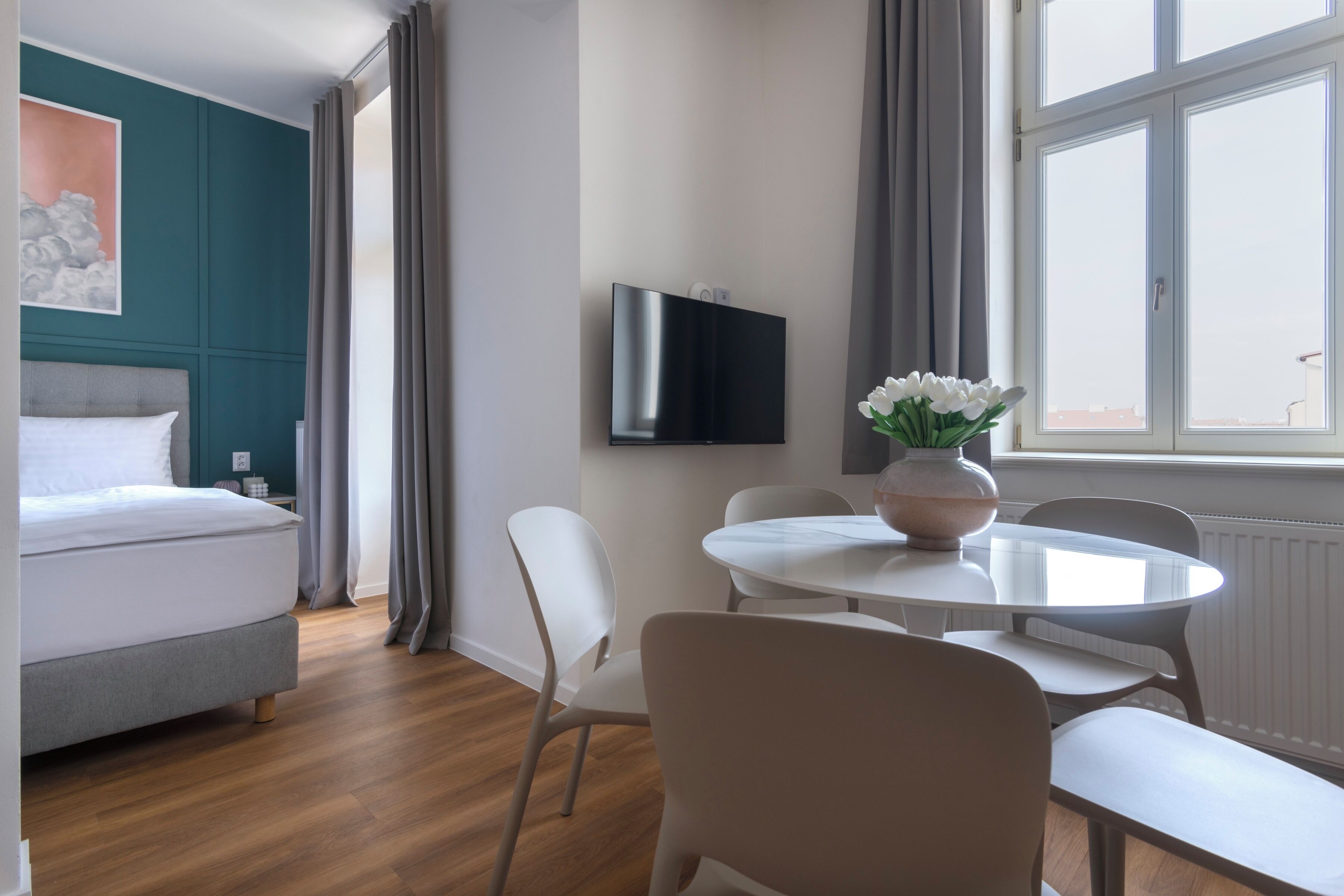 A modern hotel room with a bed, a TV mounted on the wall, a dining table with chairs, and a window with curtains.