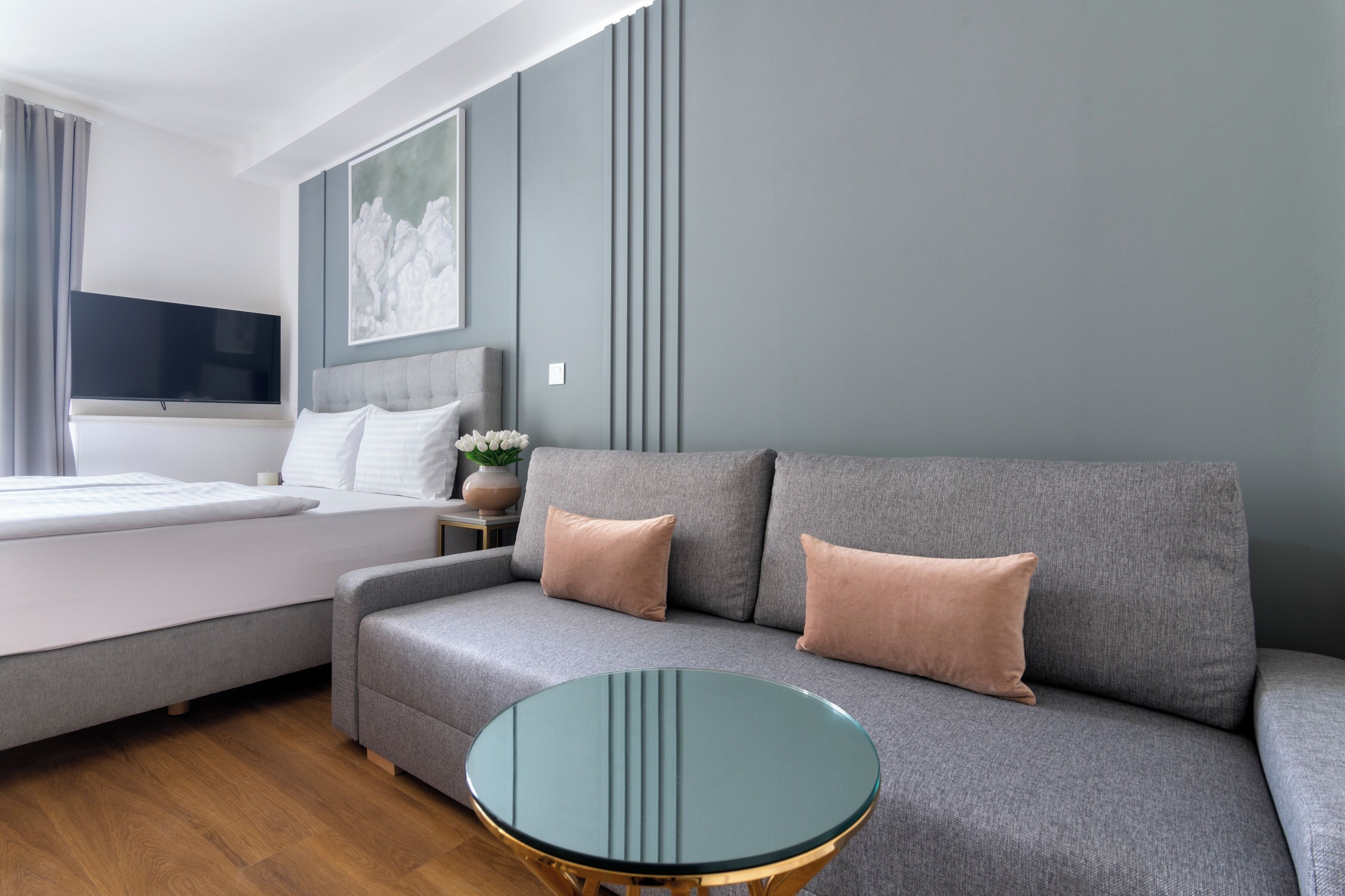 Una habitación de hotel moderna con un sofá gris, una mesa de centro de vidrio, una cama con ropa blanca y un televisor de pantalla plana montado en la pared.