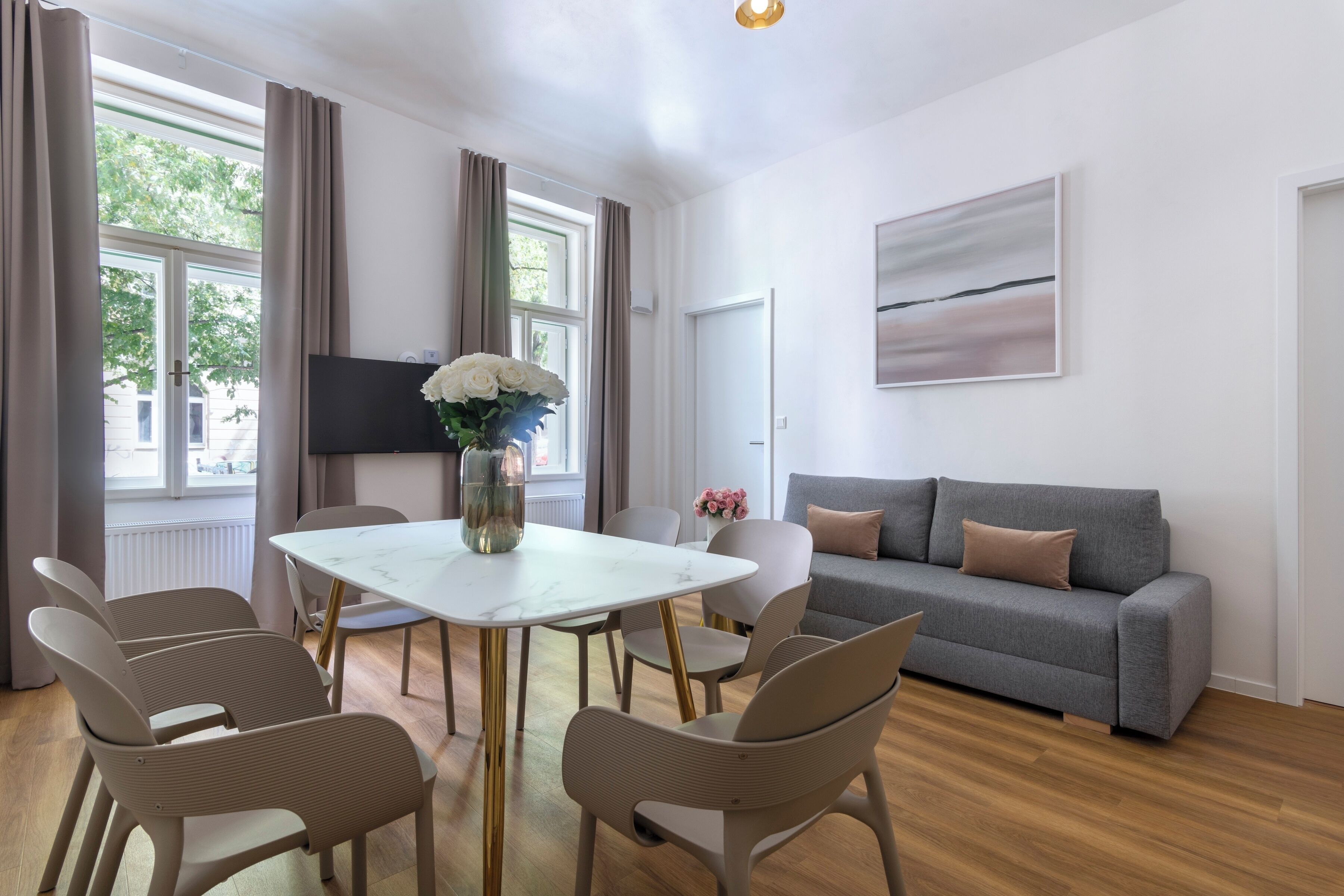 Un salon moderne avec un canapé gris, une table à manger blanche avec des chaises, un téléviseur à écran plat et un vase rempli de fleurs.