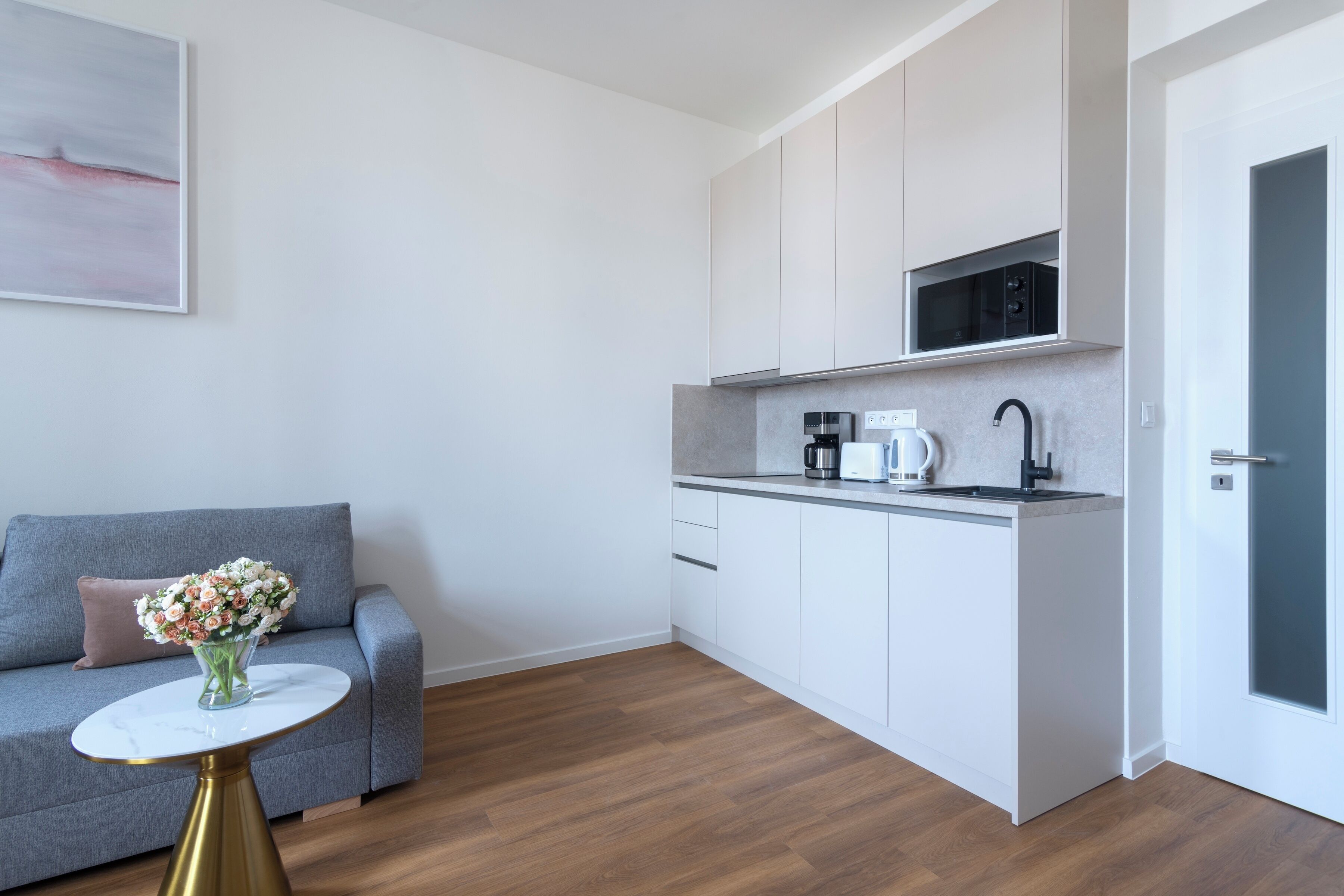 A compact kitchen with a microwave, coffee maker, and sink. There's a grey sofa with a vase of flowers and a small round table with a gold base.