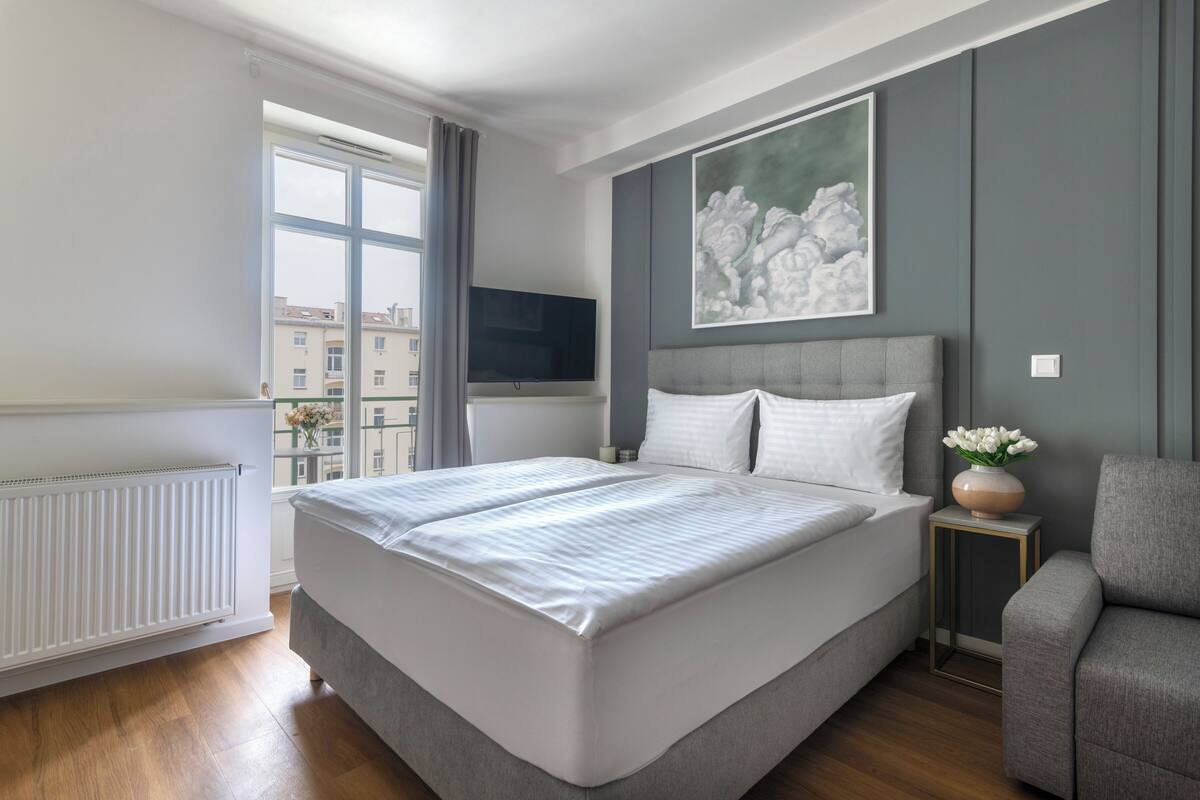 Ein modernes Schlafzimmer mit einem großen Bett, einem grauen Polsterarmstuhl, einem kleinen Tisch mit einer Vase und einem Bild an der Wand.