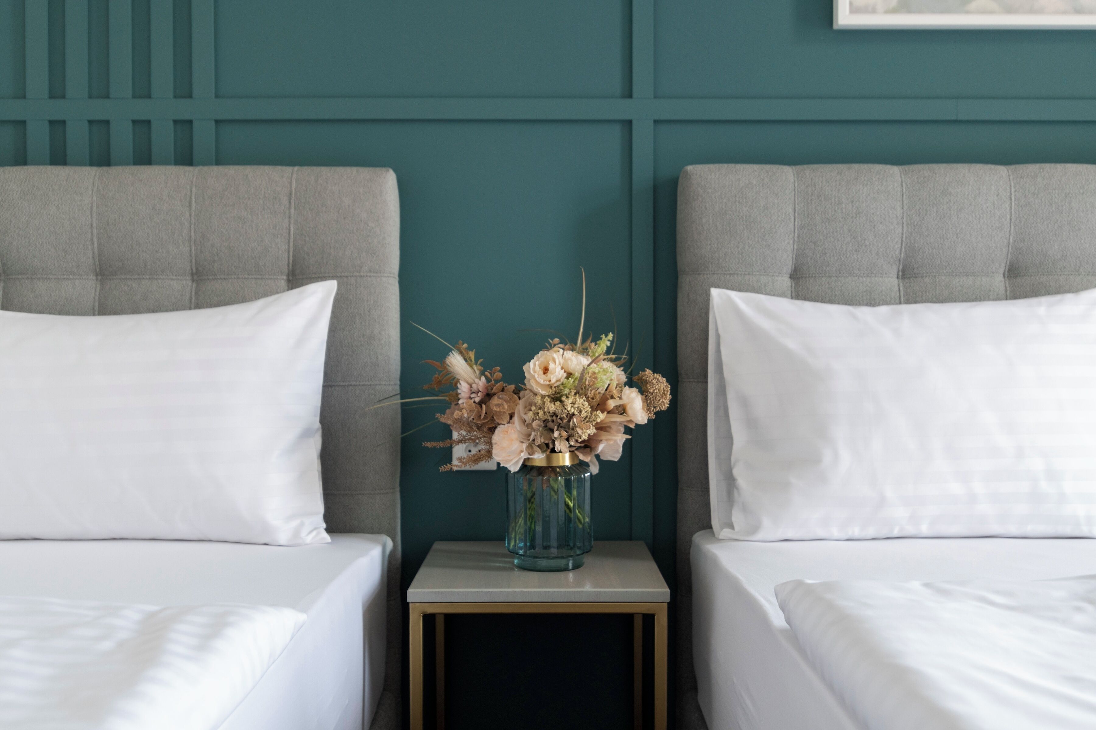 A hotel room with two beds, a small bedside table with a vase of flowers, and teal-colored walls.