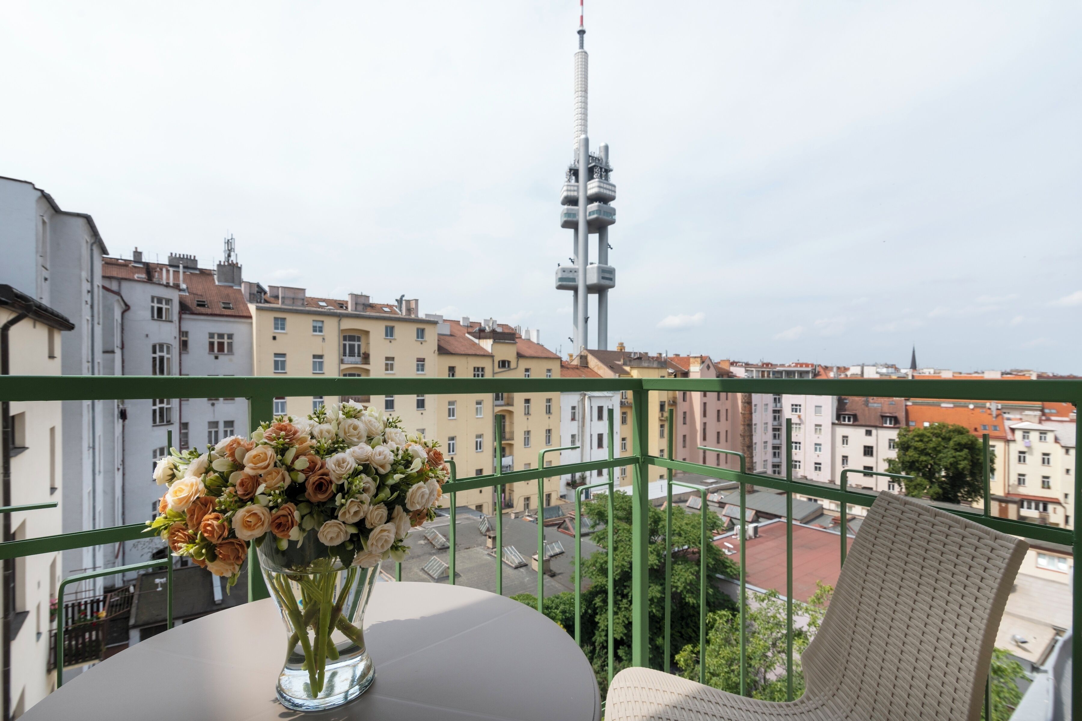 A balcony with a table and a vase of flowers, overlooking a cityscape with buildings and a tall tower.