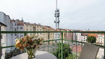 A balcony with a table and a vase of flowers, overlooking a cityscape with buildings and a tall tower.