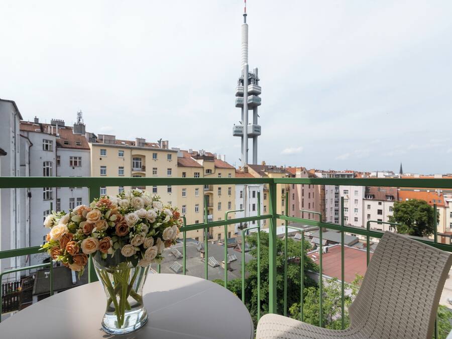 A balcony with a table and a vase of flowers, overlooking a cityscape with buildings and a tall tower.