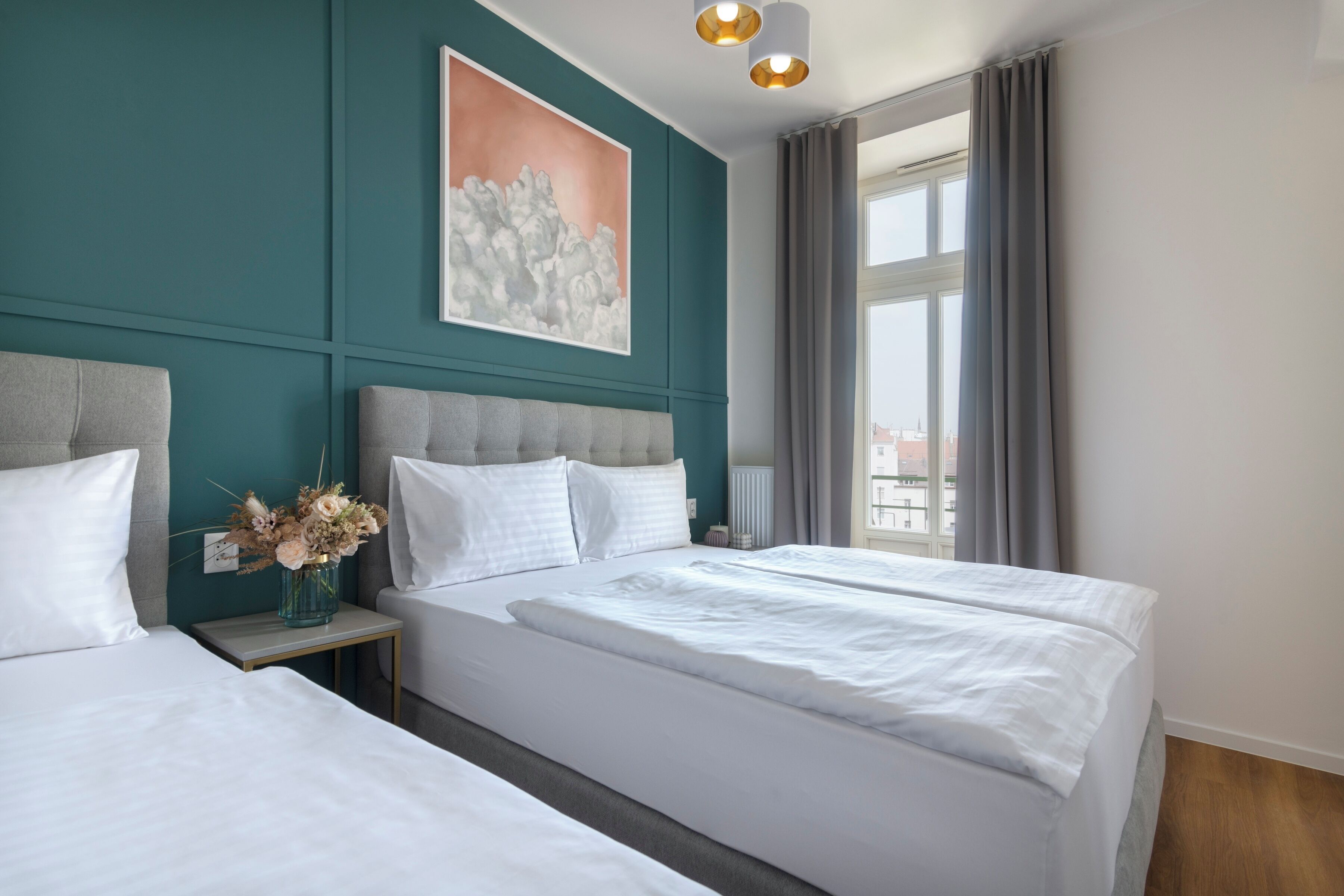 A hotel room with two beds, a large window with curtains, a painting on the wall, and a vase with flowers on the nightstand.