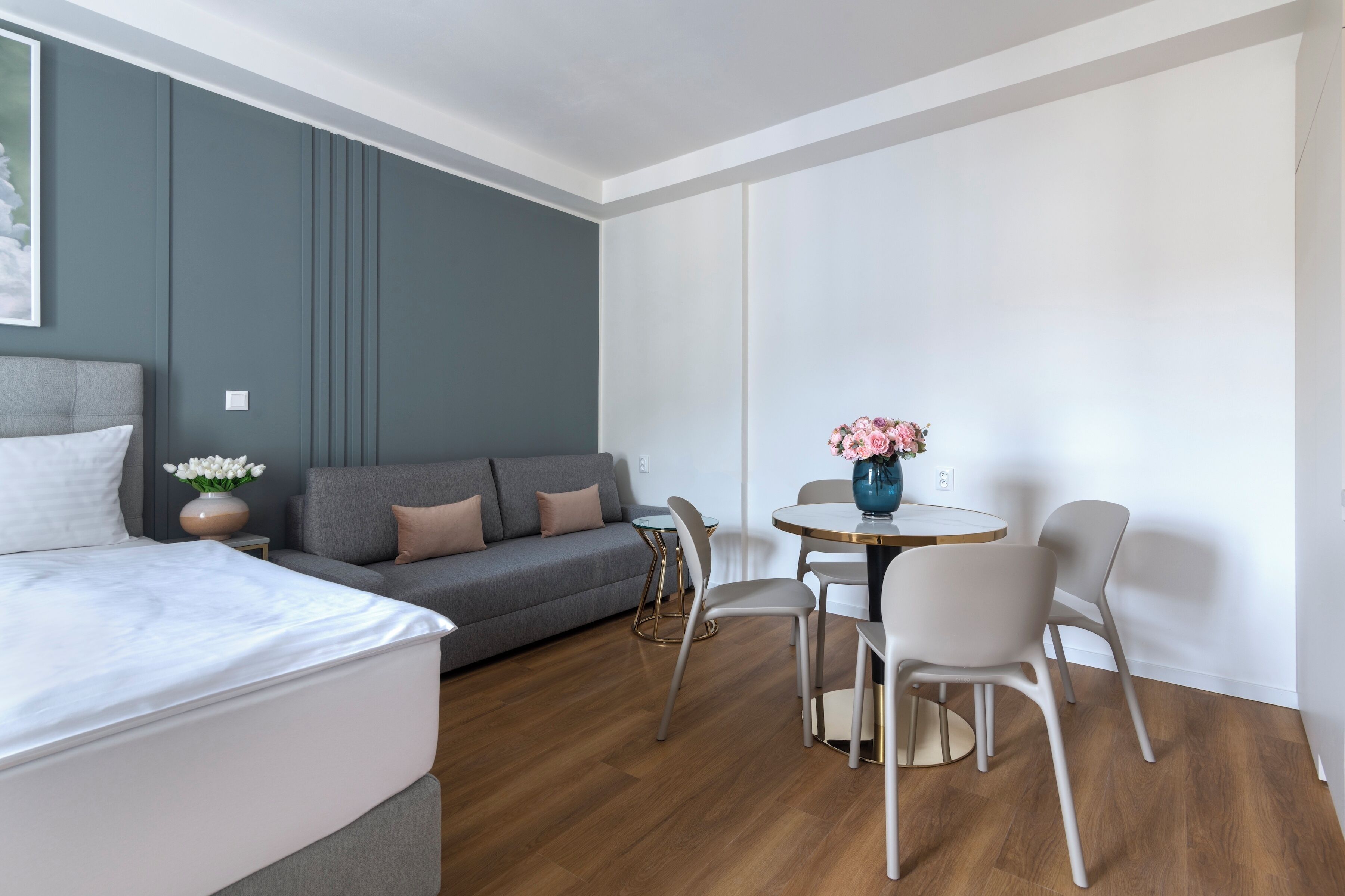 Een moderne hotelkamer met een bed, grijze bank, eettafel met stoelen, en een bloevas.