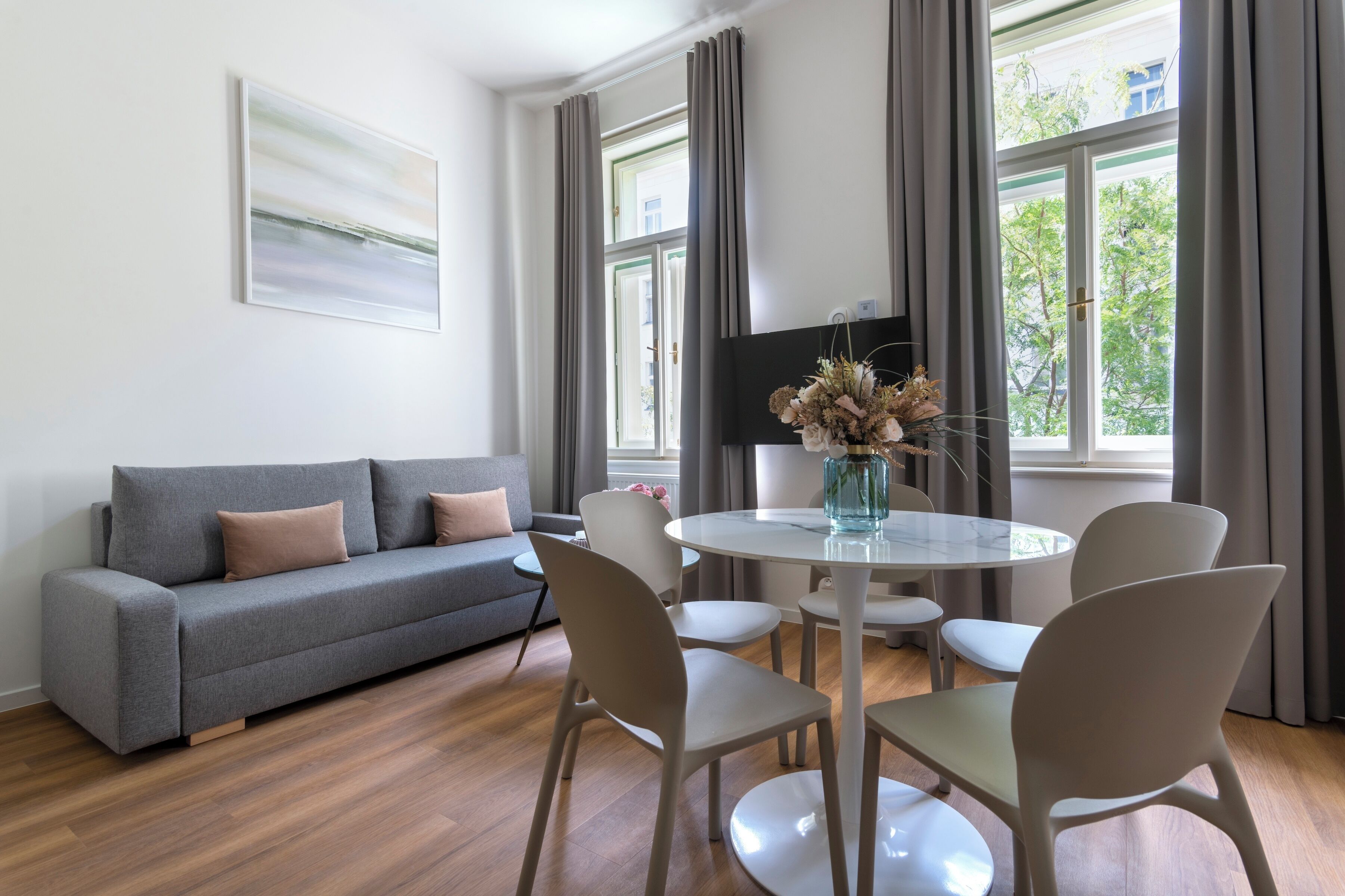 Een moderne woonkamer met een grijze bank, witte stoelen, een ronde tafel en een vaas met gedroogde bloemen.