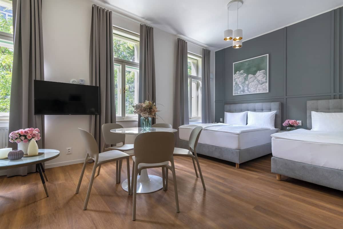 Ein modernes Hotelzimmer mit Flachbildfernseher, Essecke mit Tisch und Stühlen, zwei Betten und großen Fenstern mit Vorhängen.
