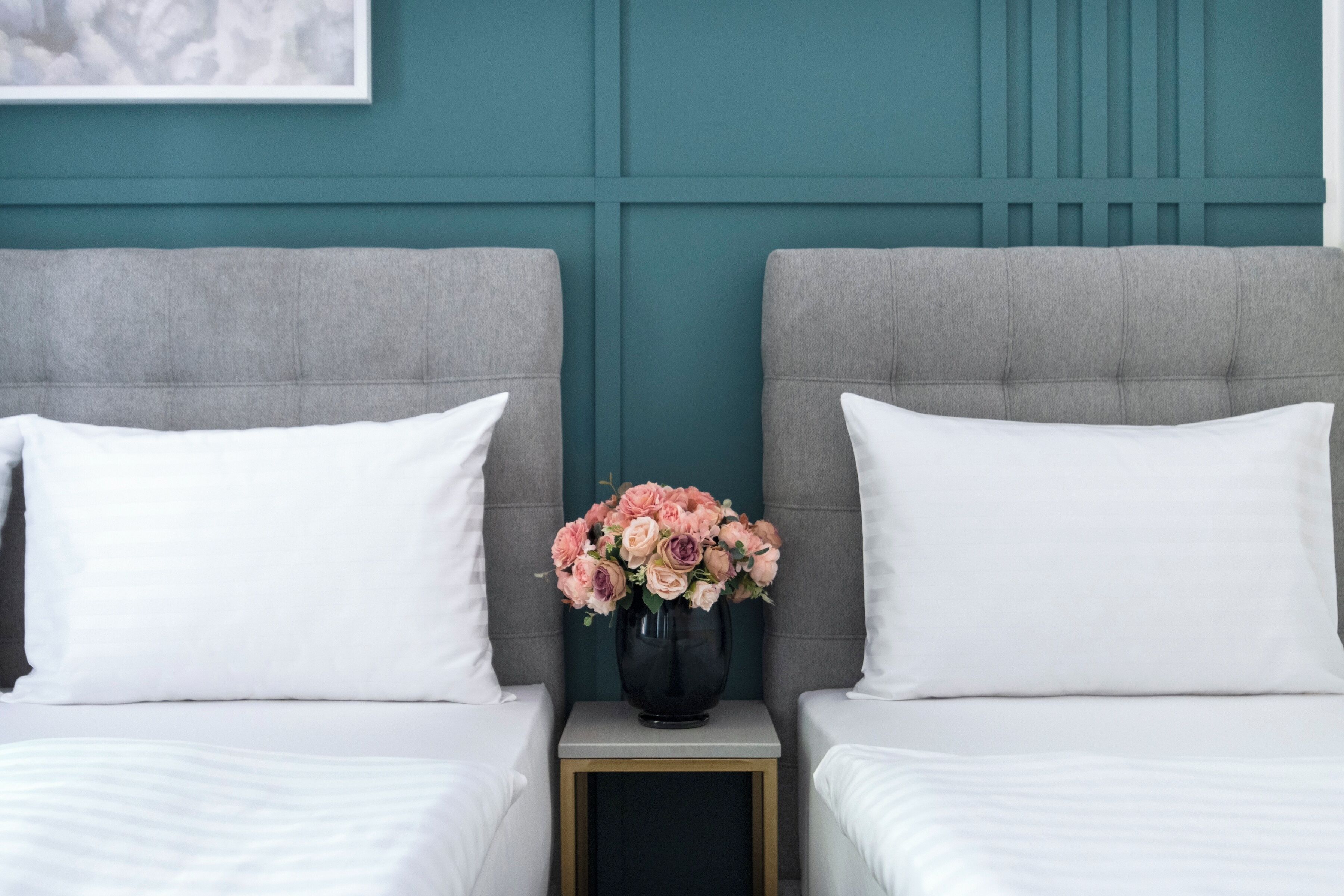 A hotel room with two beds, a nightstand with a vase of flowers, and teal-colored walls.
