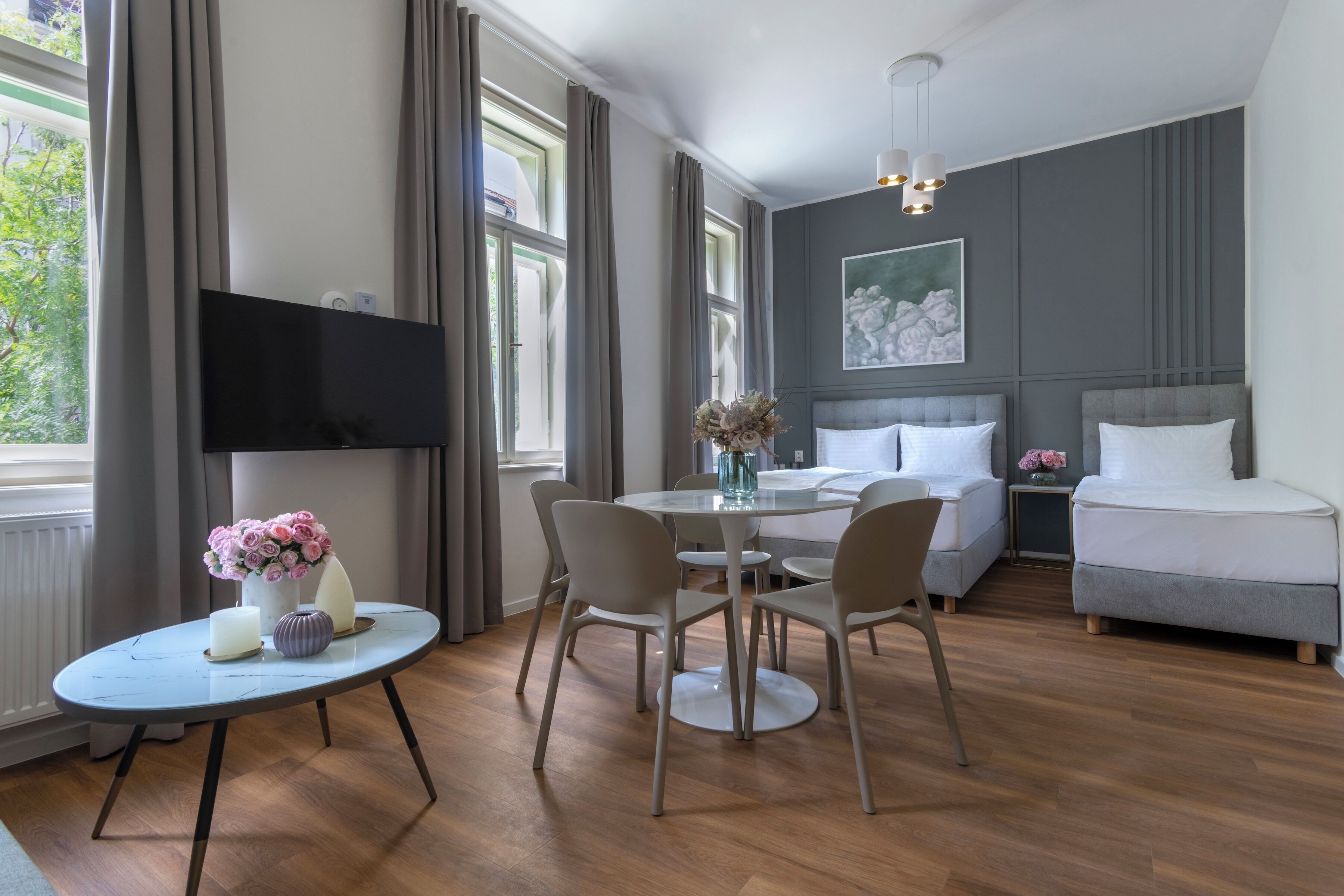 A modern hotel room with a round dining table, two sofas, a bed, and a TV mounted on the wall.