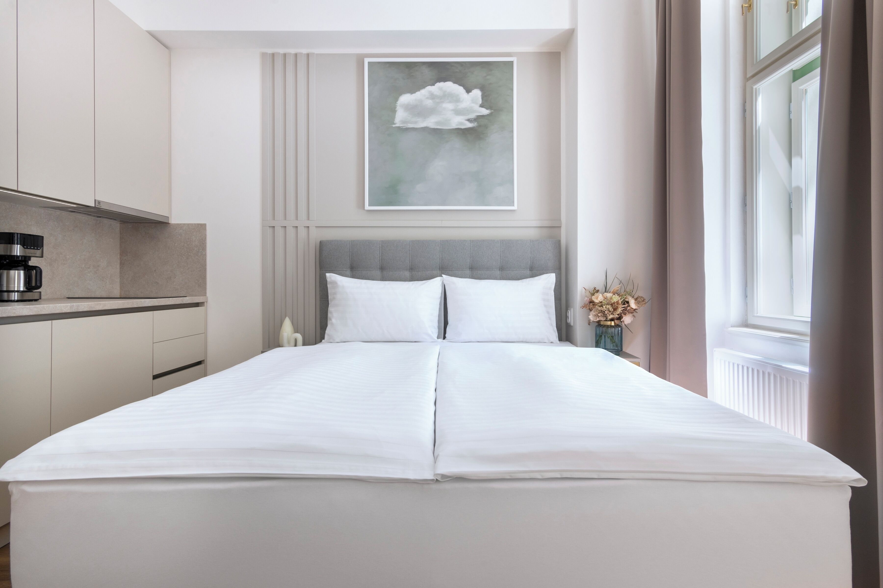 Un dormitorio moderno con una cama grande, un cabecero gris, una cafetera sobre una mesita de noche y una obra de arte enmarcada encima de la cama.