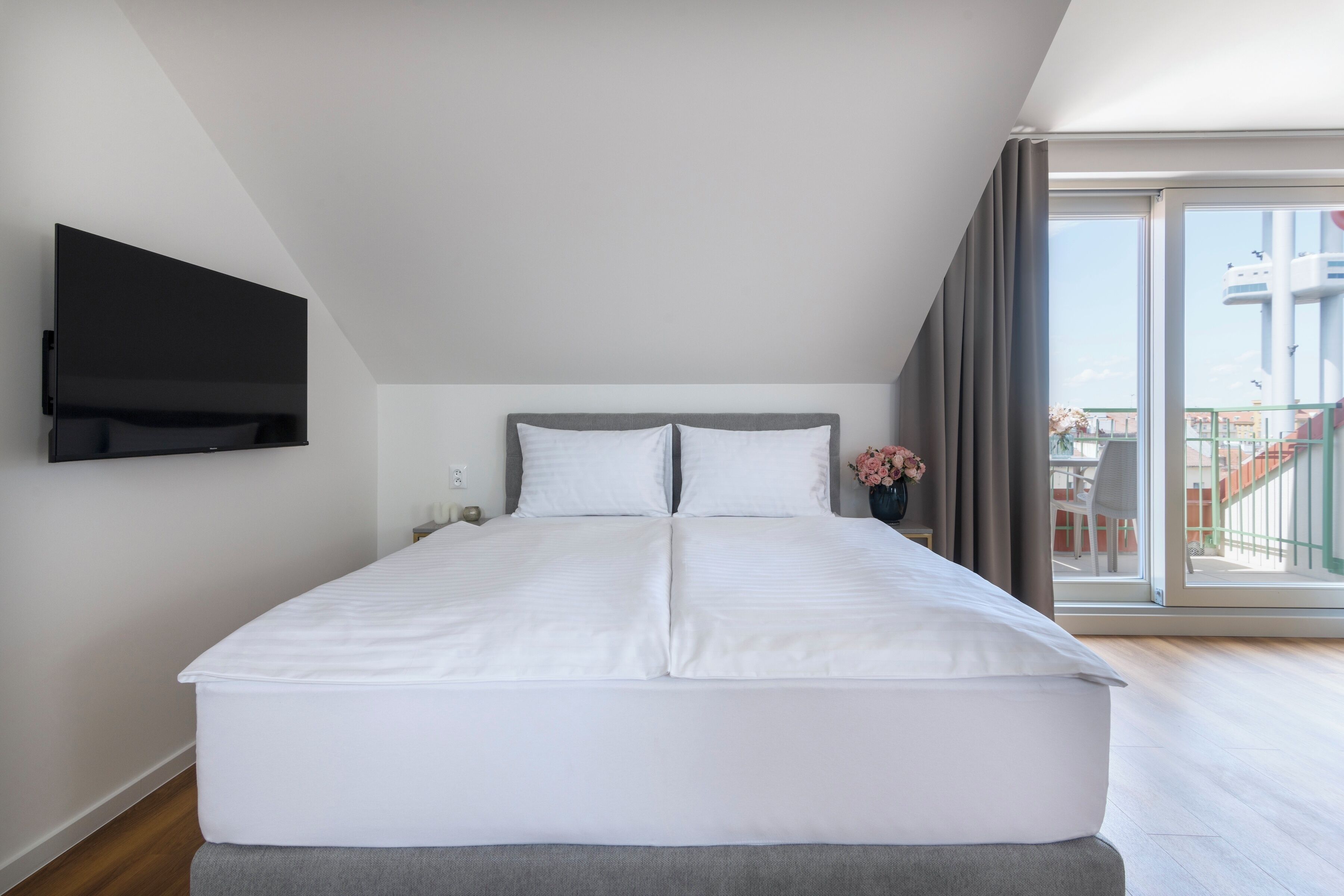 A modern bedroom with a large bed, a flat-screen TV mounted on the wall, and a balcony with a view of a building.