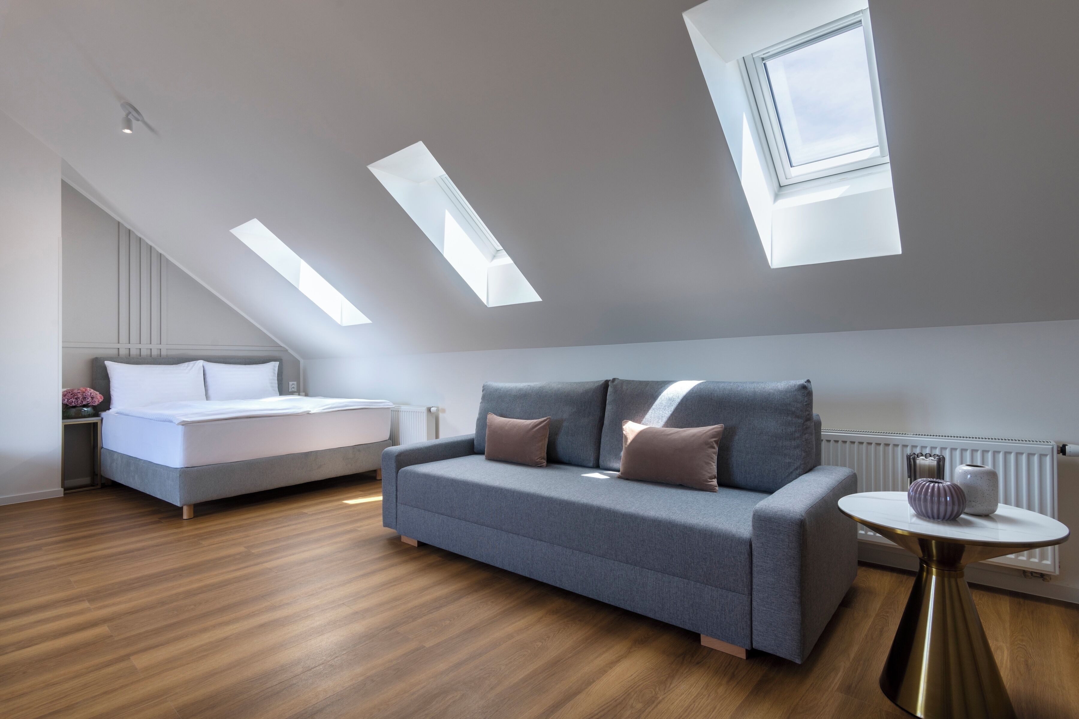 A modern loft bedroom with a sloped ceiling, skylights, a bed, a sofa, and a small table.