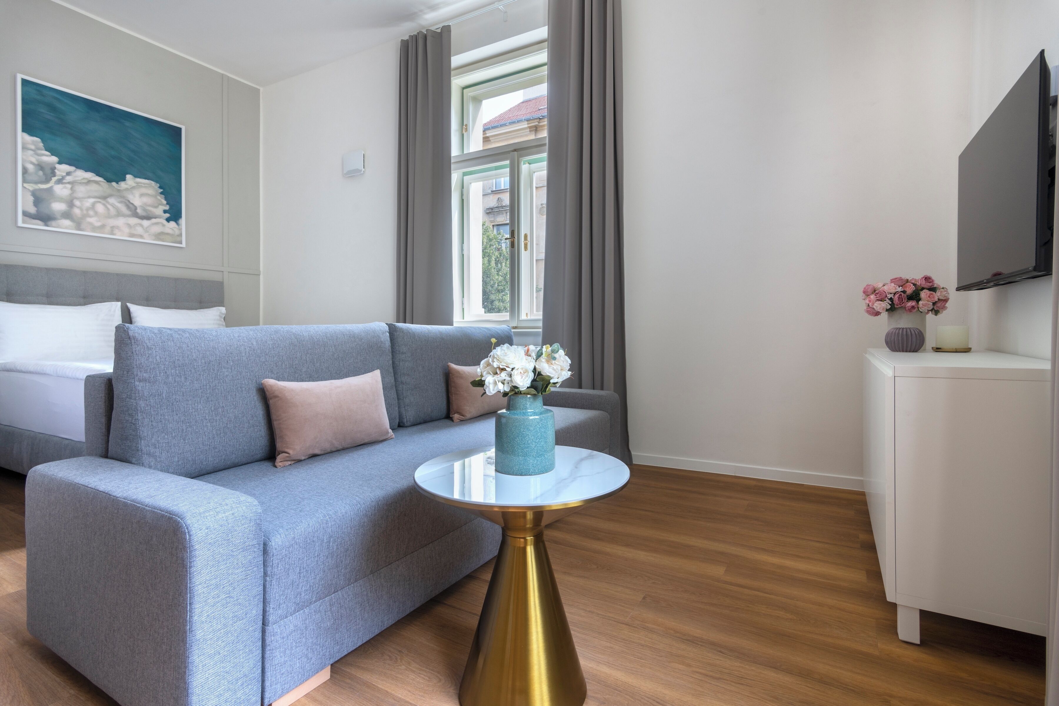 Een moderne hotelkamer met een grijze bank, een ronde gouden tafel en een televisie aan de muur.