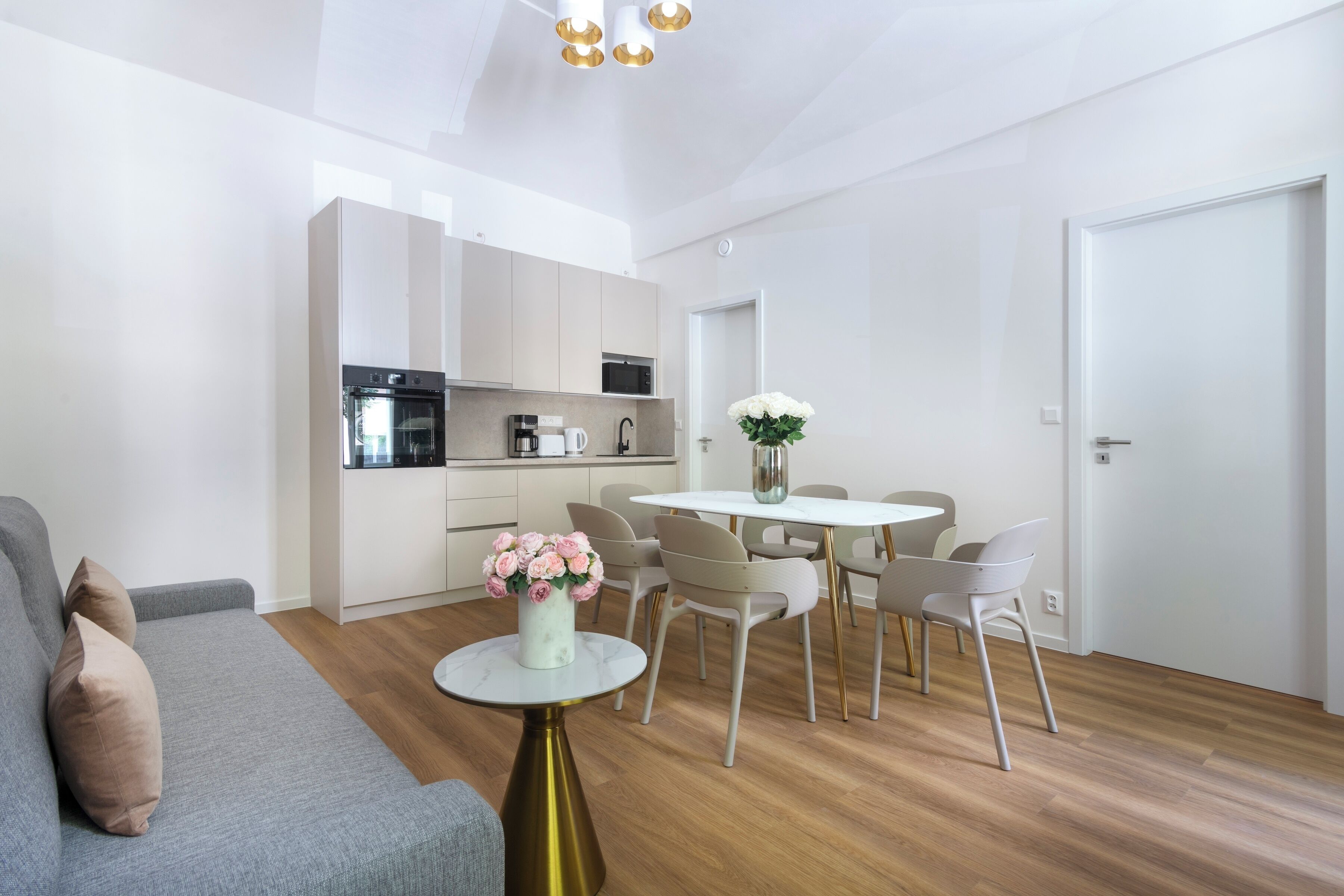 Una sala moderna con un sofá gris, una mesita redonda con un florero y una zona de comedor con una mesa y sillas blancas.