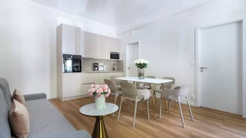 Una sala moderna con un sofá gris, una mesita redonda con un florero y una zona de comedor con una mesa y sillas blancas.